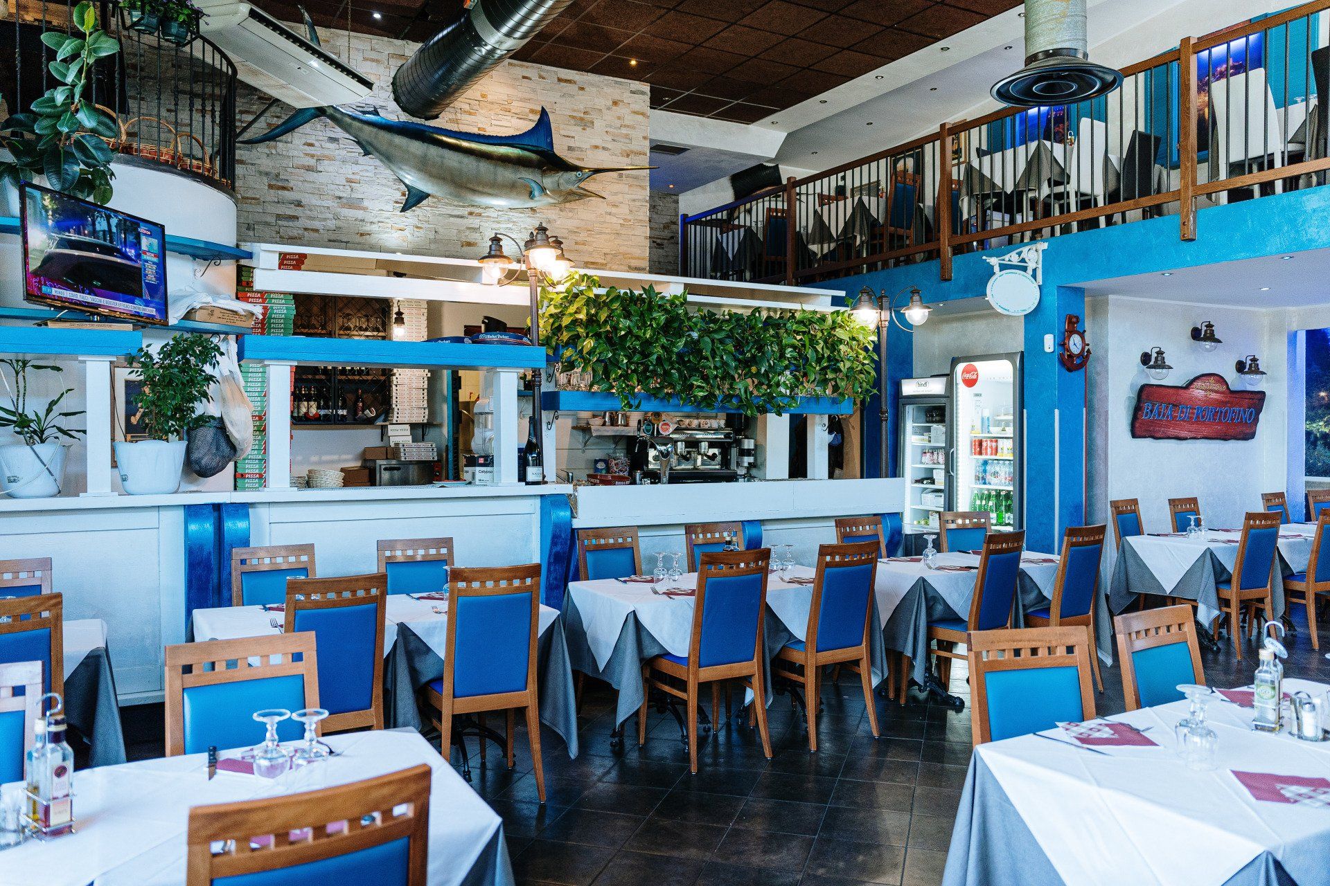 Interno di un ristorante greco con decorazioni blu e bianche, tavoli apparecchiati e zona bar.