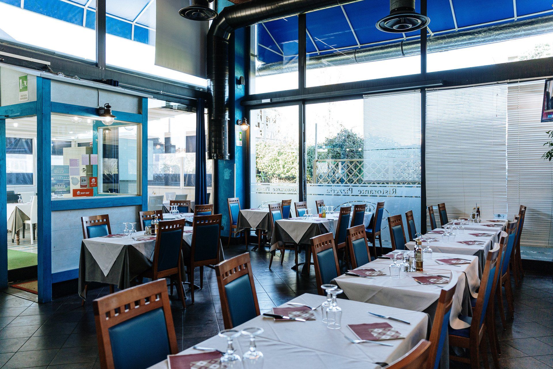 Interno del ristorante con tavoli apparecchiati per la cena; decorazioni blu e bianche con grandi finestre.