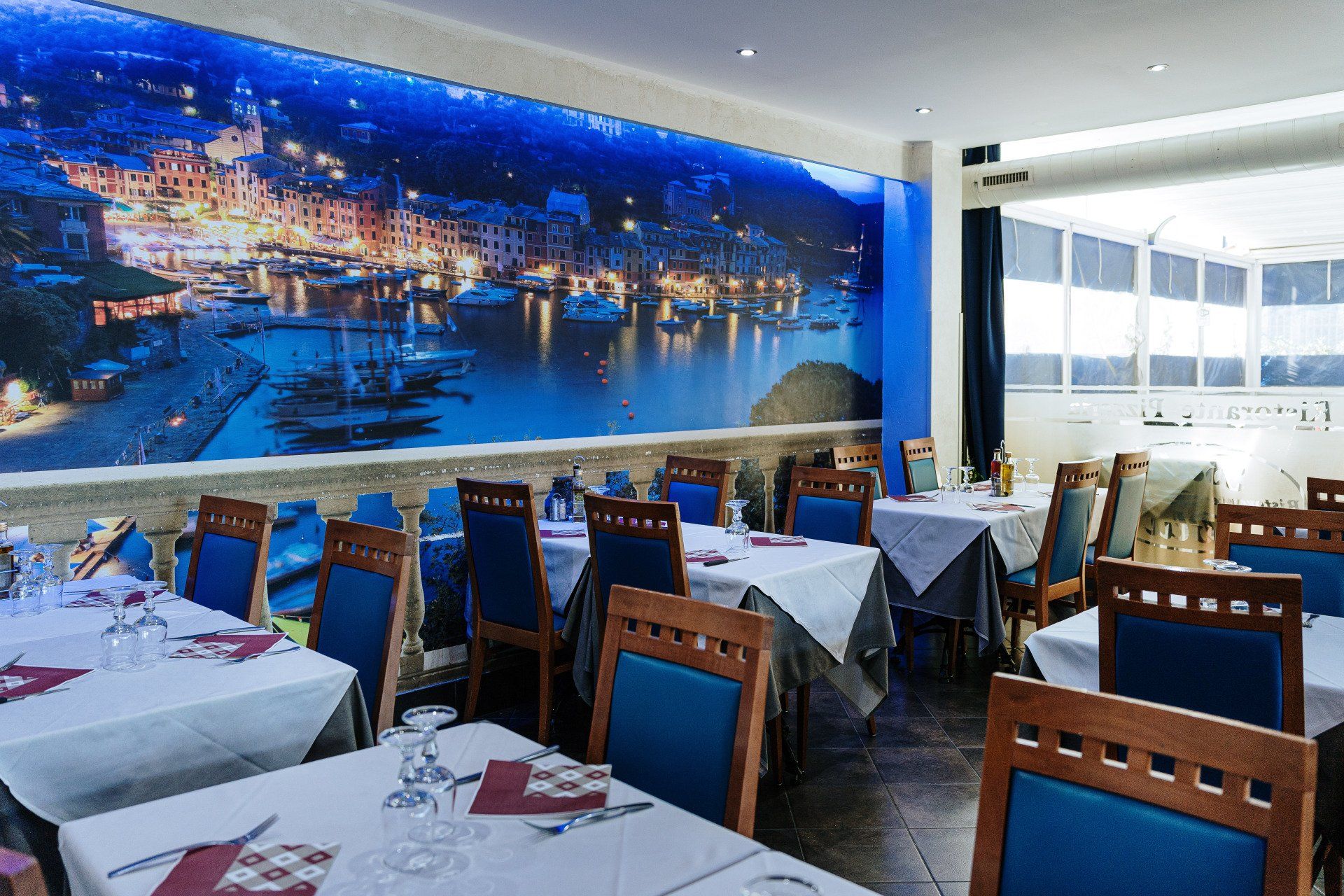 Interno del ristorante con tavoli apparecchiati per cenare e un murale raffigurante un porto di notte.