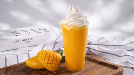 Mango smoothie topped with whipped cream beside two sliced mangoes on a wooden table