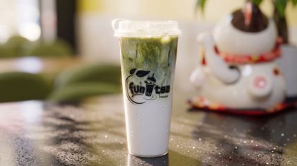 Iced green-and-white drink in a clear cup on a table, with a blurred dessert in the background.