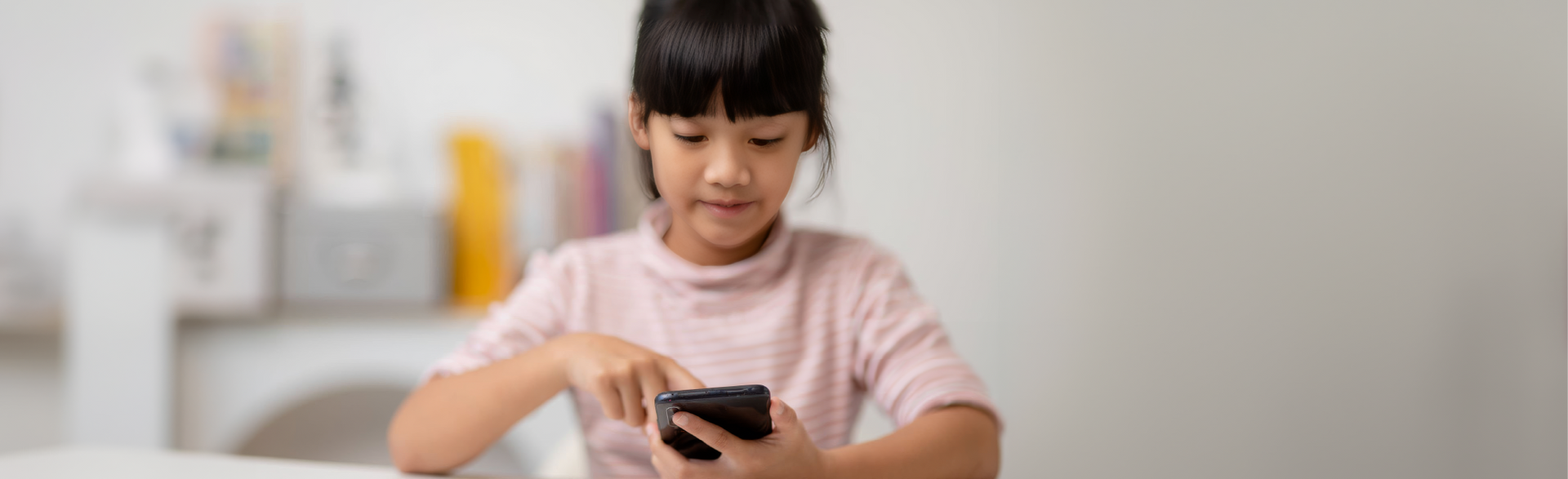 A person in a light pink shirt sitting at a table and interacting with a smartphone in a brightly lit, blurred room.