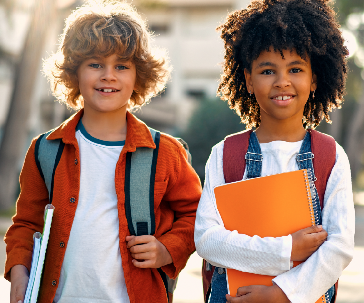 Two children with backpacks and books smiling outside.