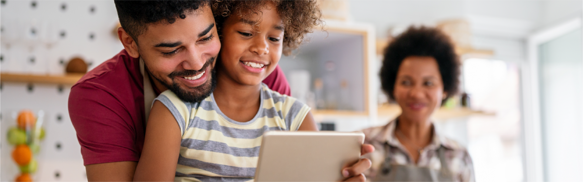 Father and daughter smile while looking at a tablet together, with mother in background.
