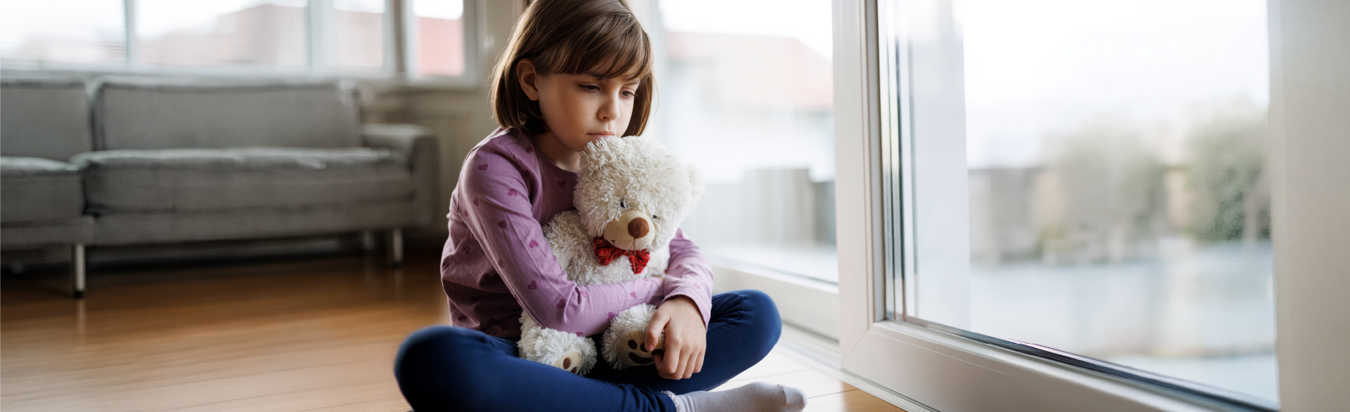 A person sitting on a floor holding a plush toy while looking out a window.