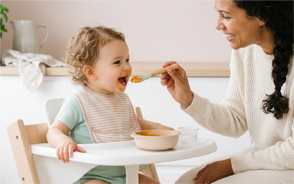 Baby being fed with a spoon, wearing a white bib and looking up with mouth open.