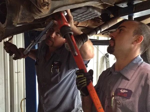 Two mechanics working under a car, using tools. One looks up, one tightens.