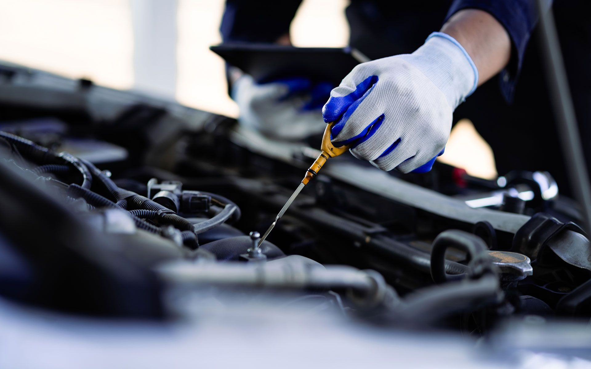Mechanic checking engine oil level with dipstick, wearing gloves, holding a tablet.