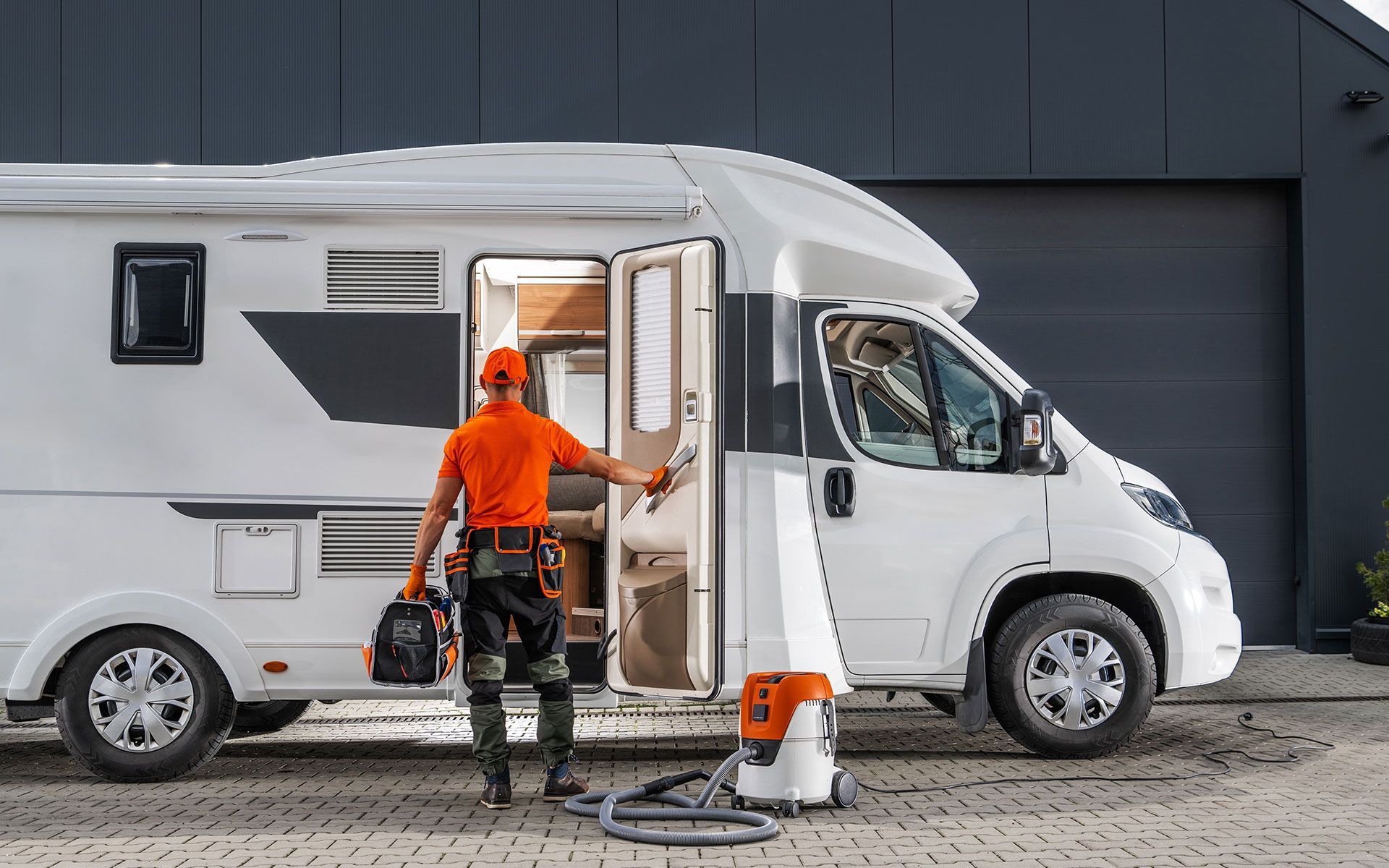 Man in orange shirt with tools stands by a white RV, next to a vacuum.