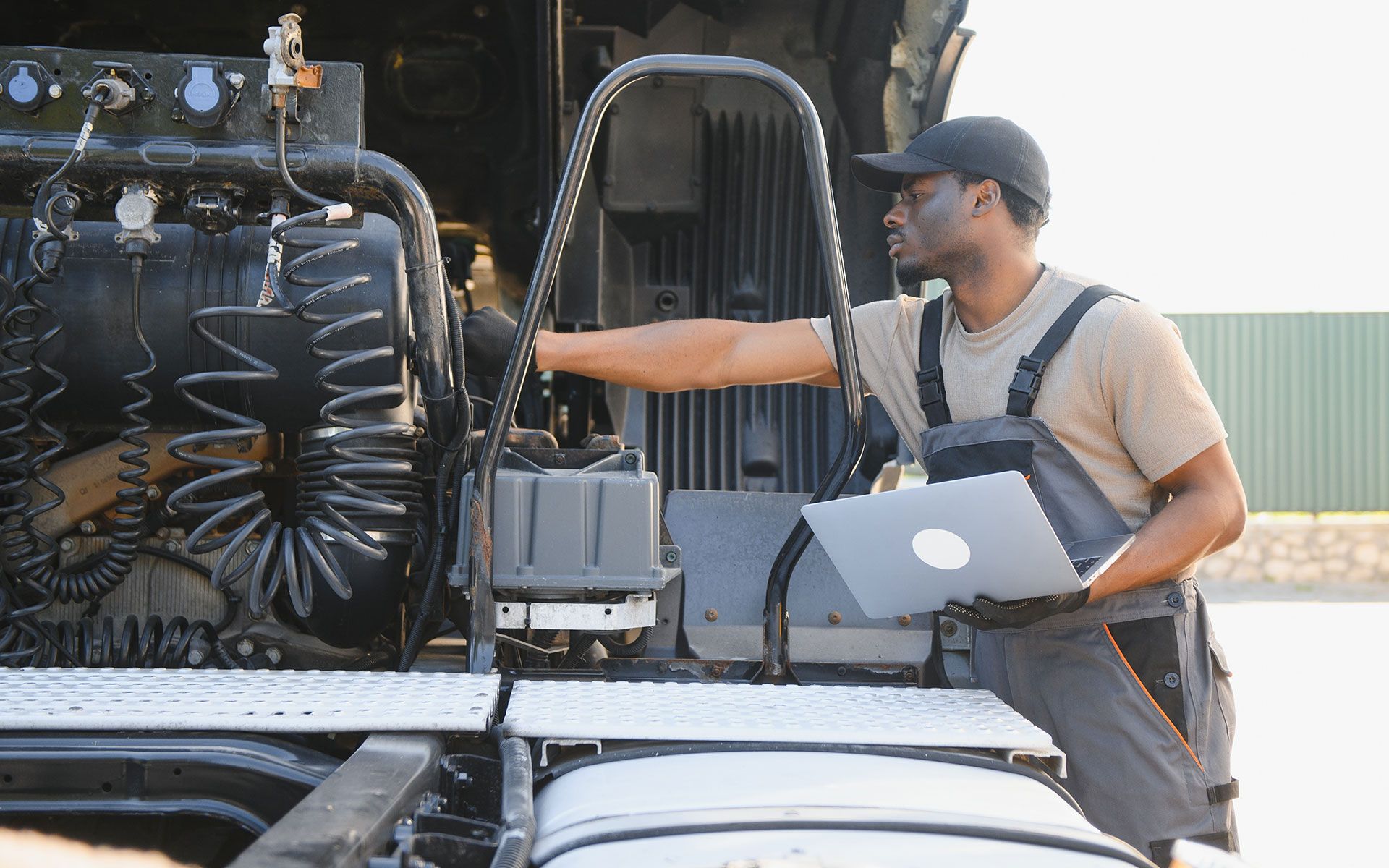 Mechanic inspecting a truck engine, holding a laptop outdoors.