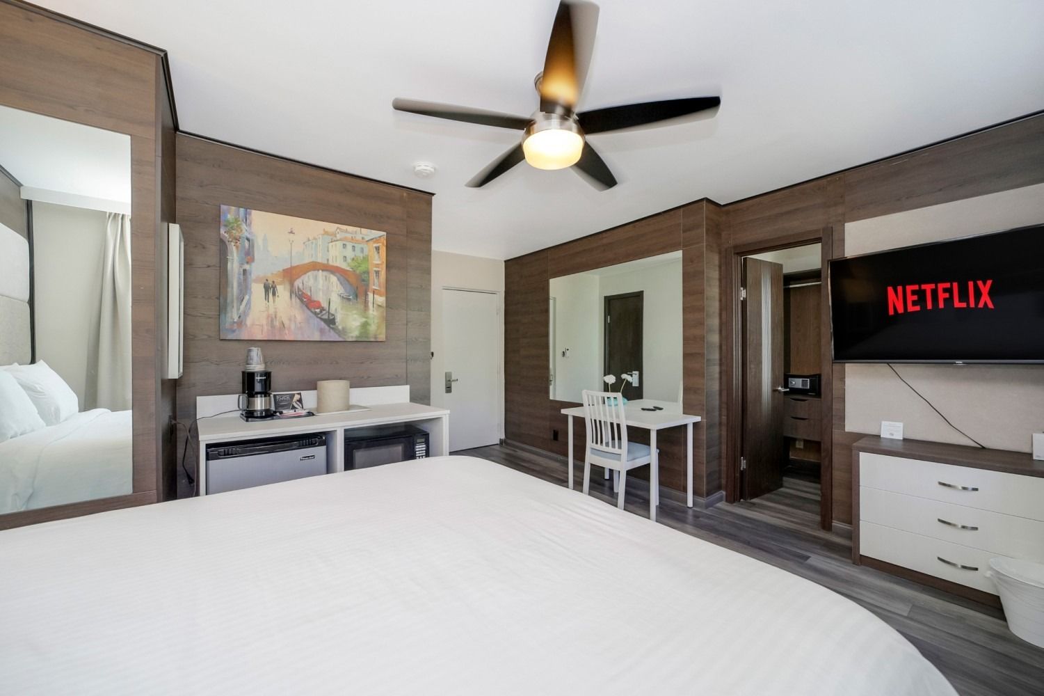 Hotel room with king-size bed, TV with Netflix, desk, artwork, coffee maker, mirror. Brown and white decor.