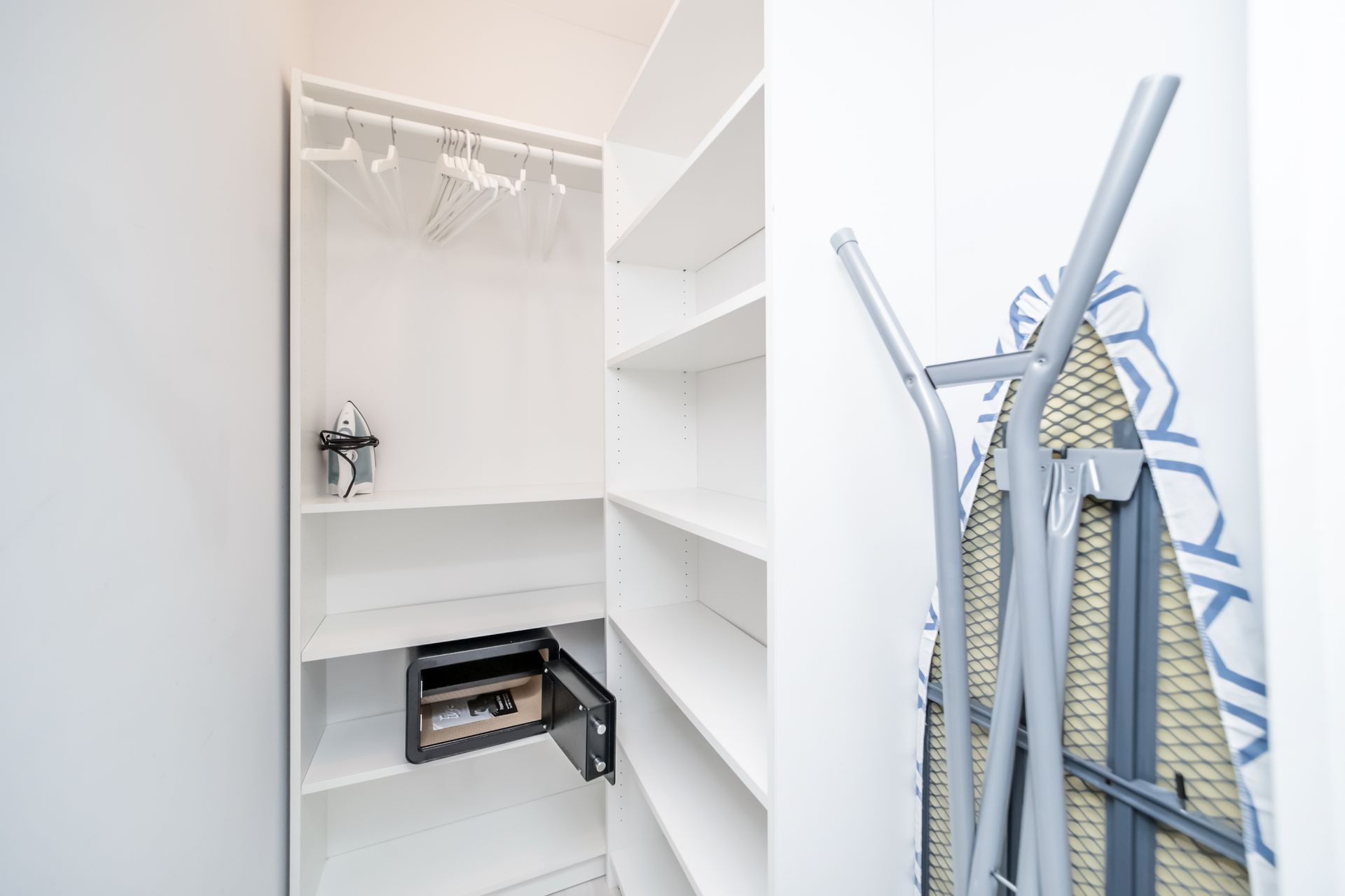 White closet with shelves, hangers, safe, and folded ironing board against a wall.