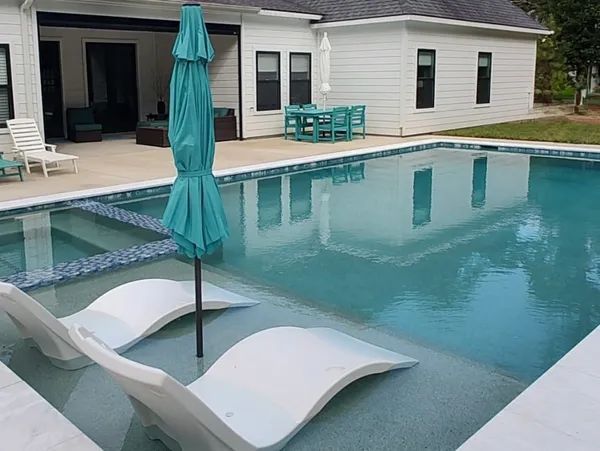 Modern backyard pool with lounge chairs, teal umbrella, and a white patio beside a house