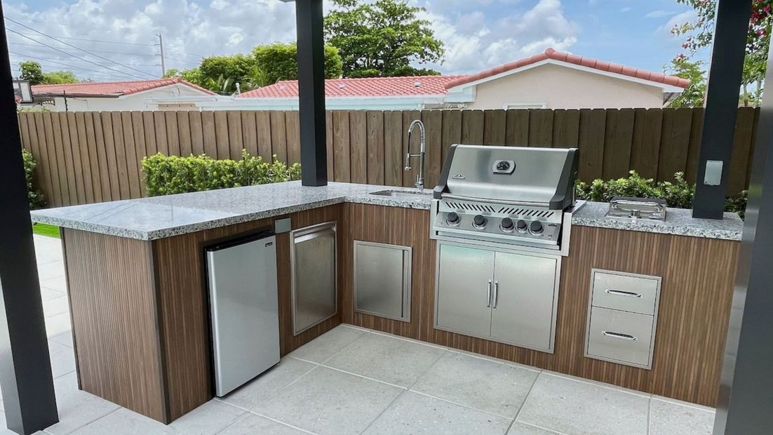 Outdoor patio kitchen with white countertops, wood cabinets, sink, grill, and mini fridge.