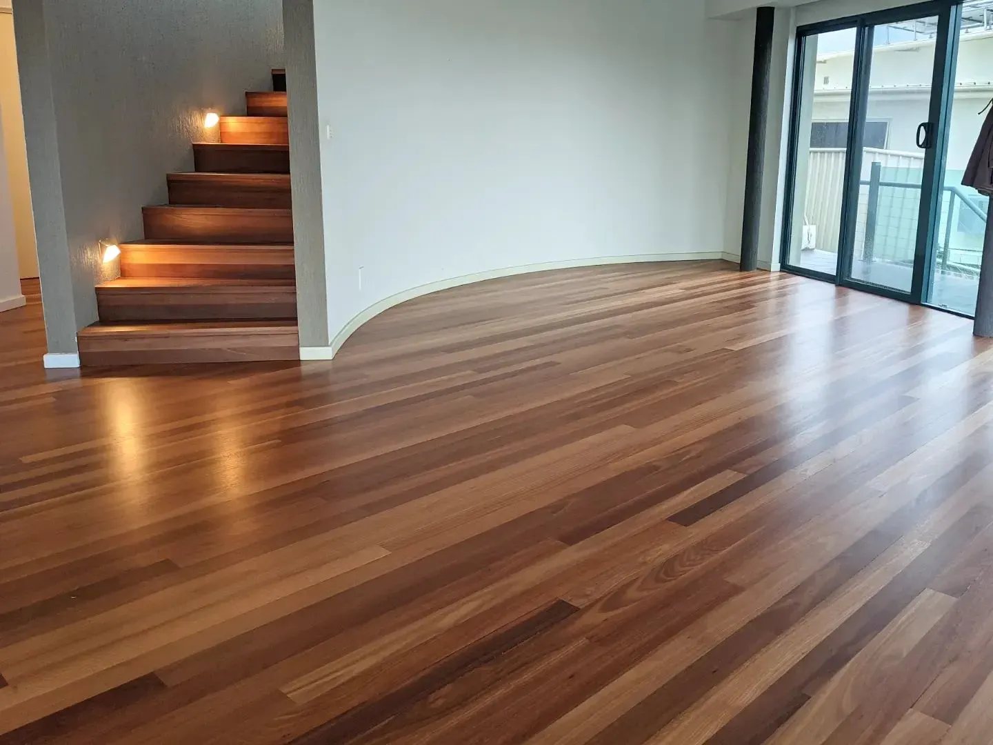 Empty modern living room with wood floors, curved wall, lit staircase, and large sliding glass doors