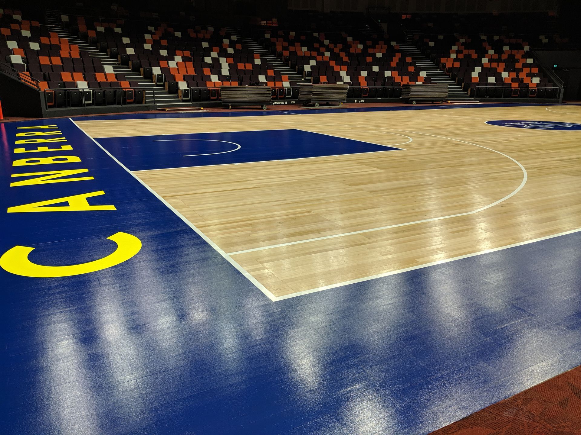 Empty indoor basketball court with blue and wood flooring and orange seats in the background