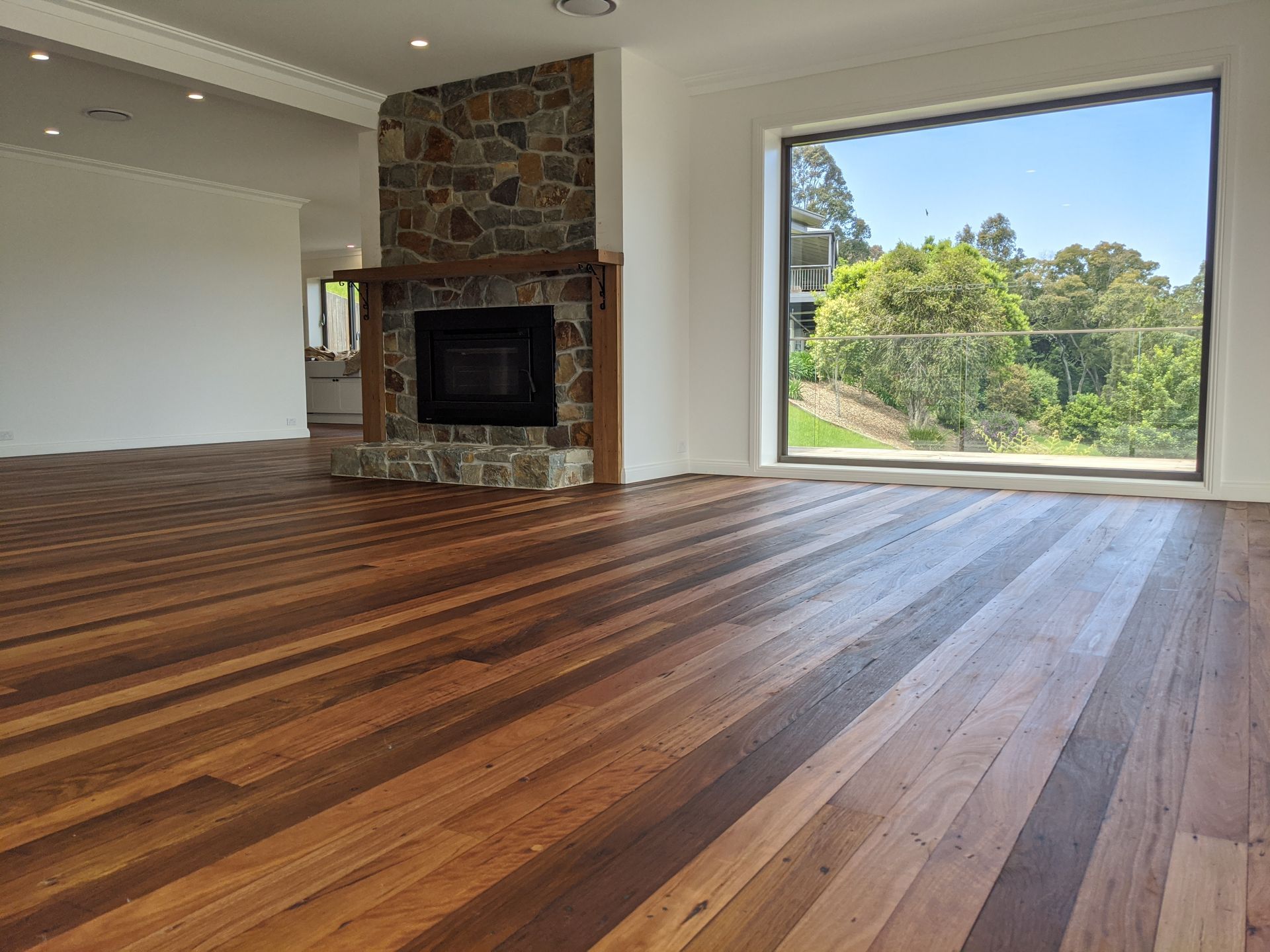 Empty modern living room with hardwood floors, stone fireplace, and a large window overlooking trees