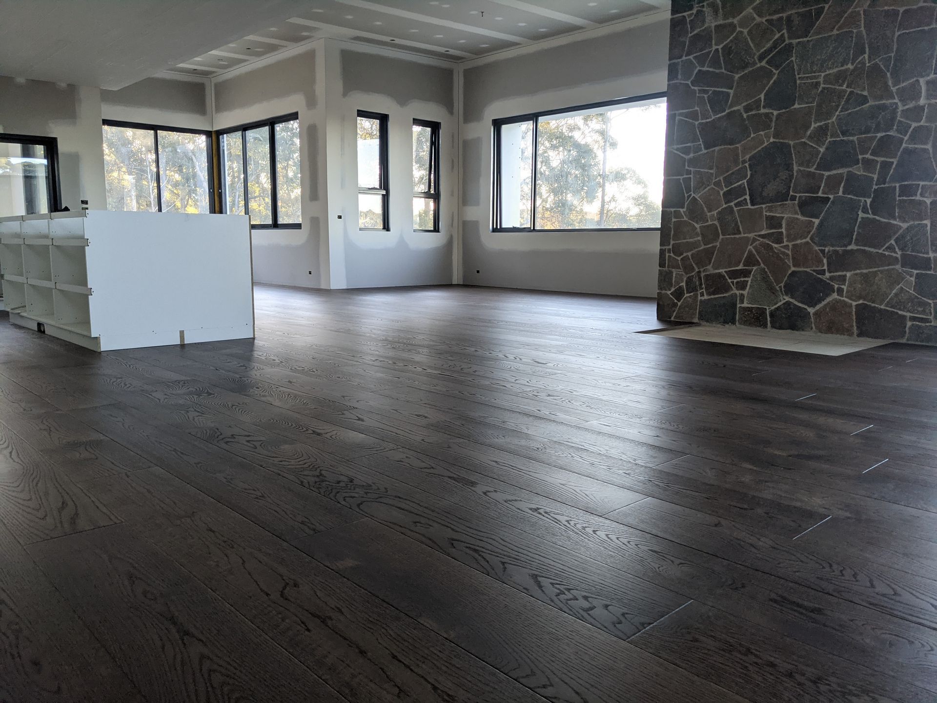 Empty modern living room with dark wood floors, large windows, and a stone fireplace