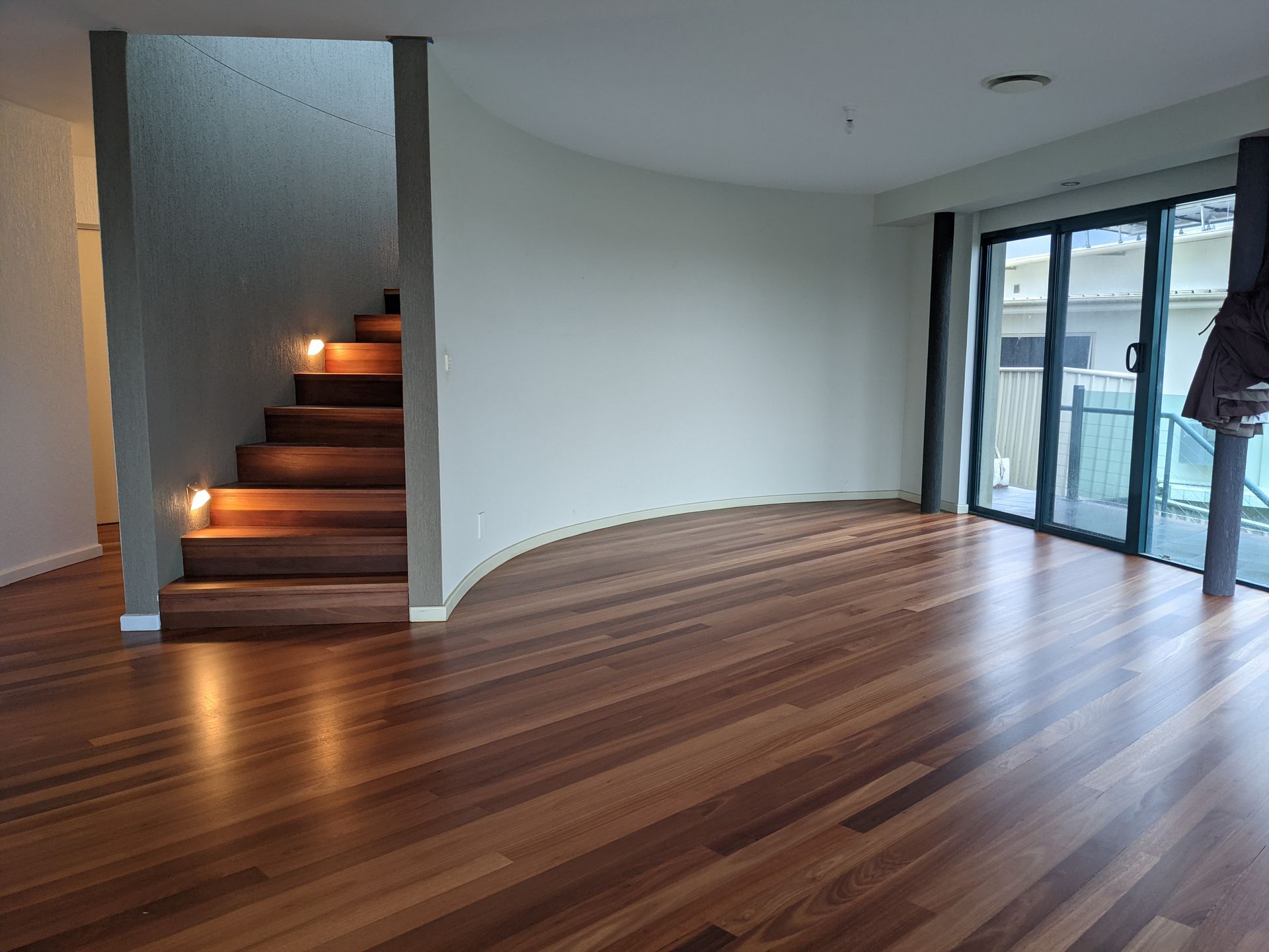Empty modern living room with polished wood floors, curved white wall, and lit stairs beside sliding glass doors