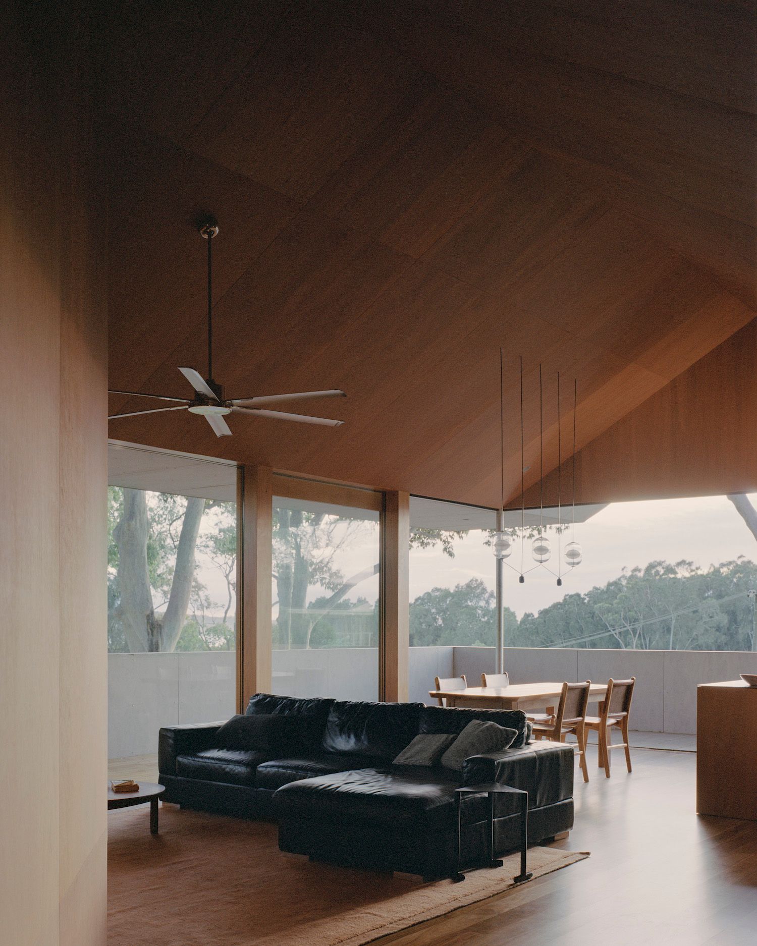 Modern living room with large windows, wood ceiling, black sectional sofa, dining table, and ceiling fan