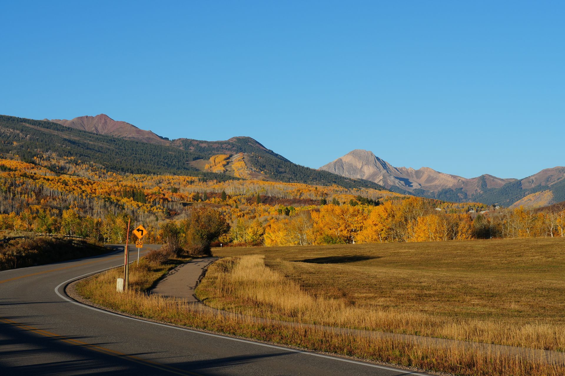 Curving road through golden autumn fields with mountains under a clear blue sky  with slow & say hello in colorado