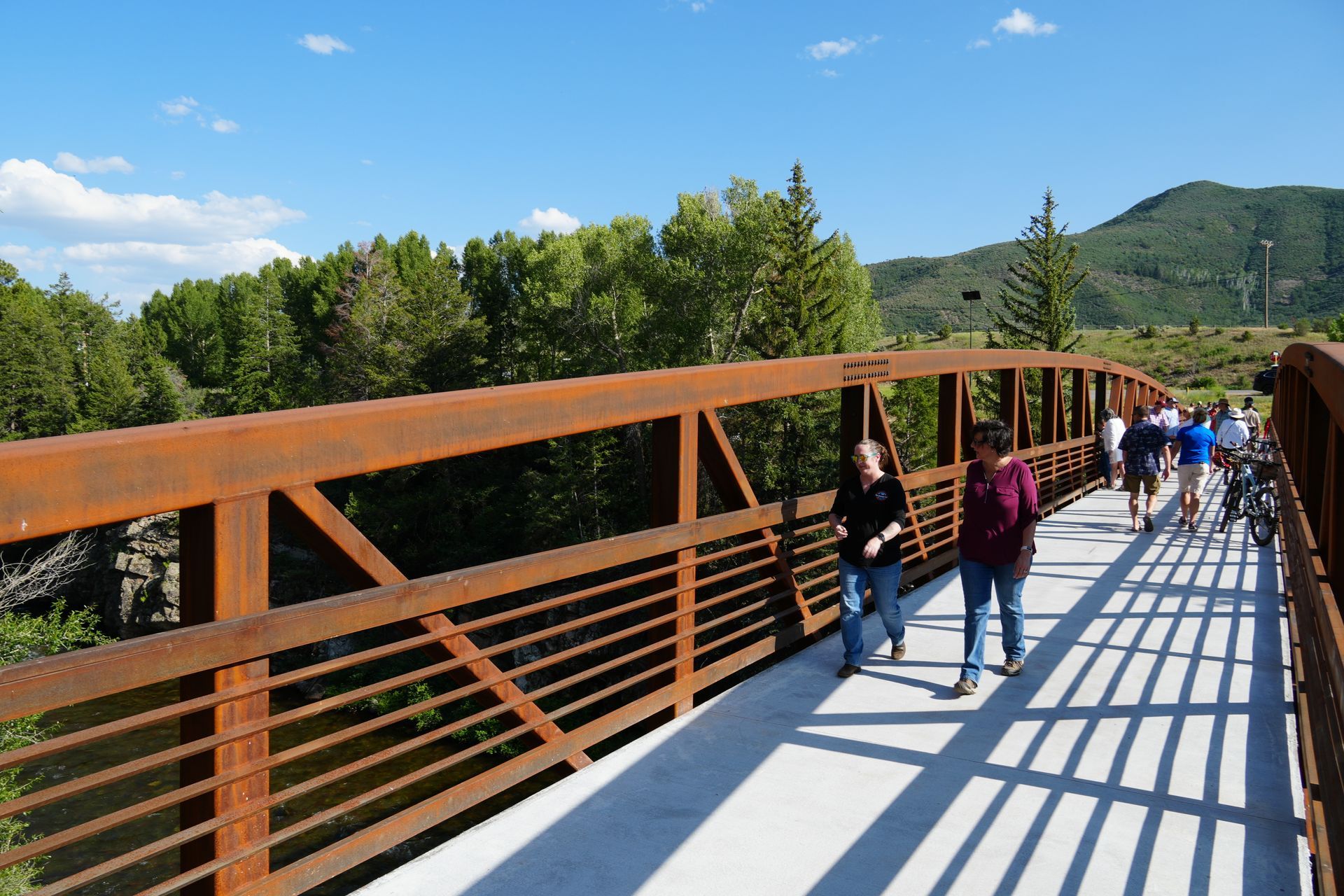People walking on a sunlit bridge with orange railings, surrounded by green hills and trees with slow & say hello in colorado
