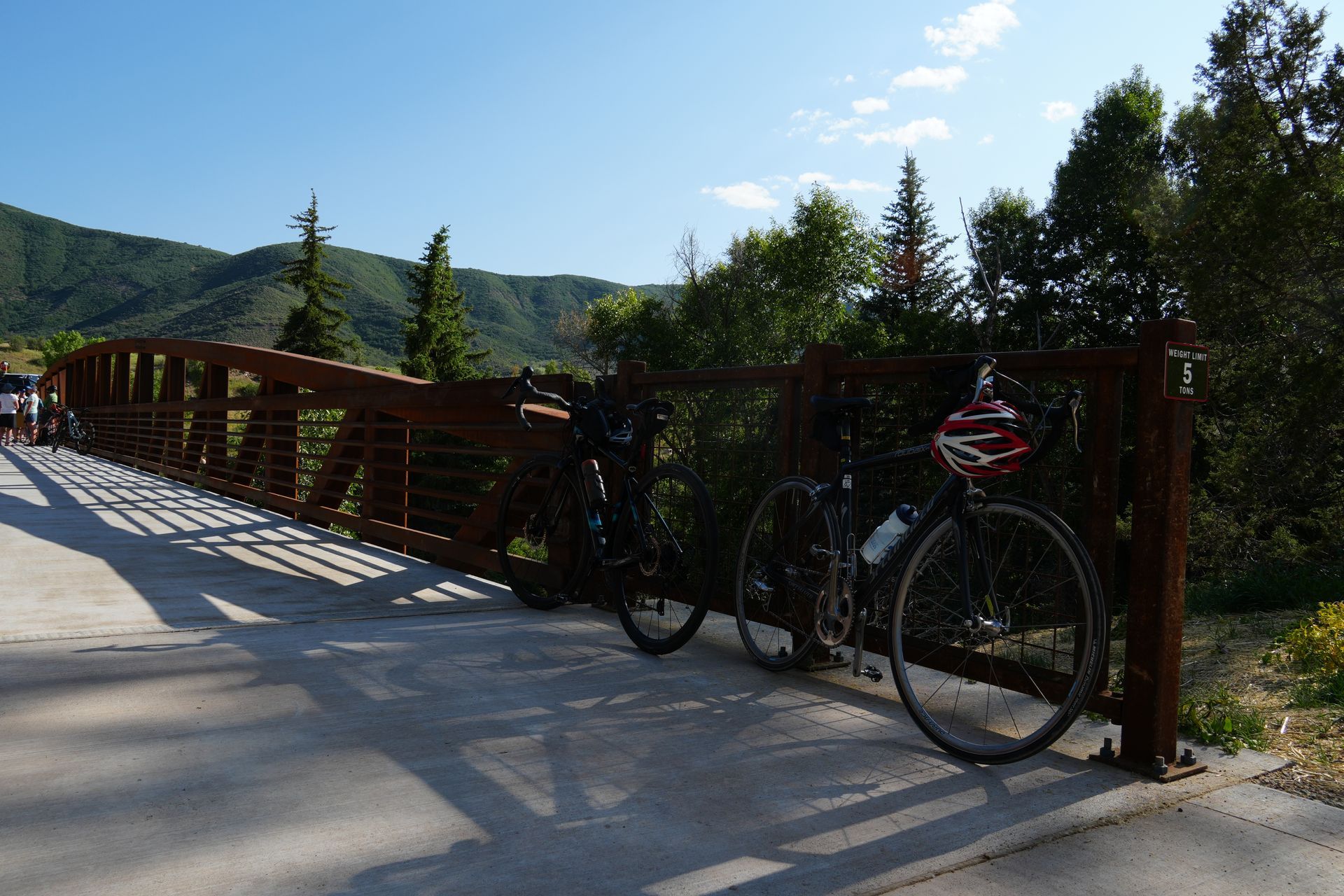 Bike on a red bridge beside a sunny mountain trail, with trees and hills in the background  with slow & say hello in colorado