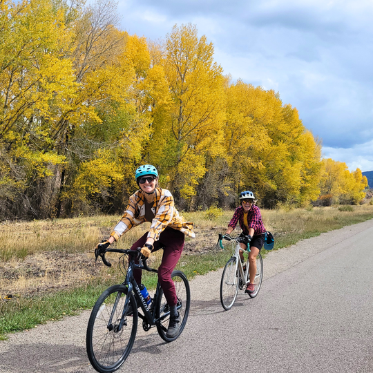 two cyclists riding on a paved path with yellow trees in the background