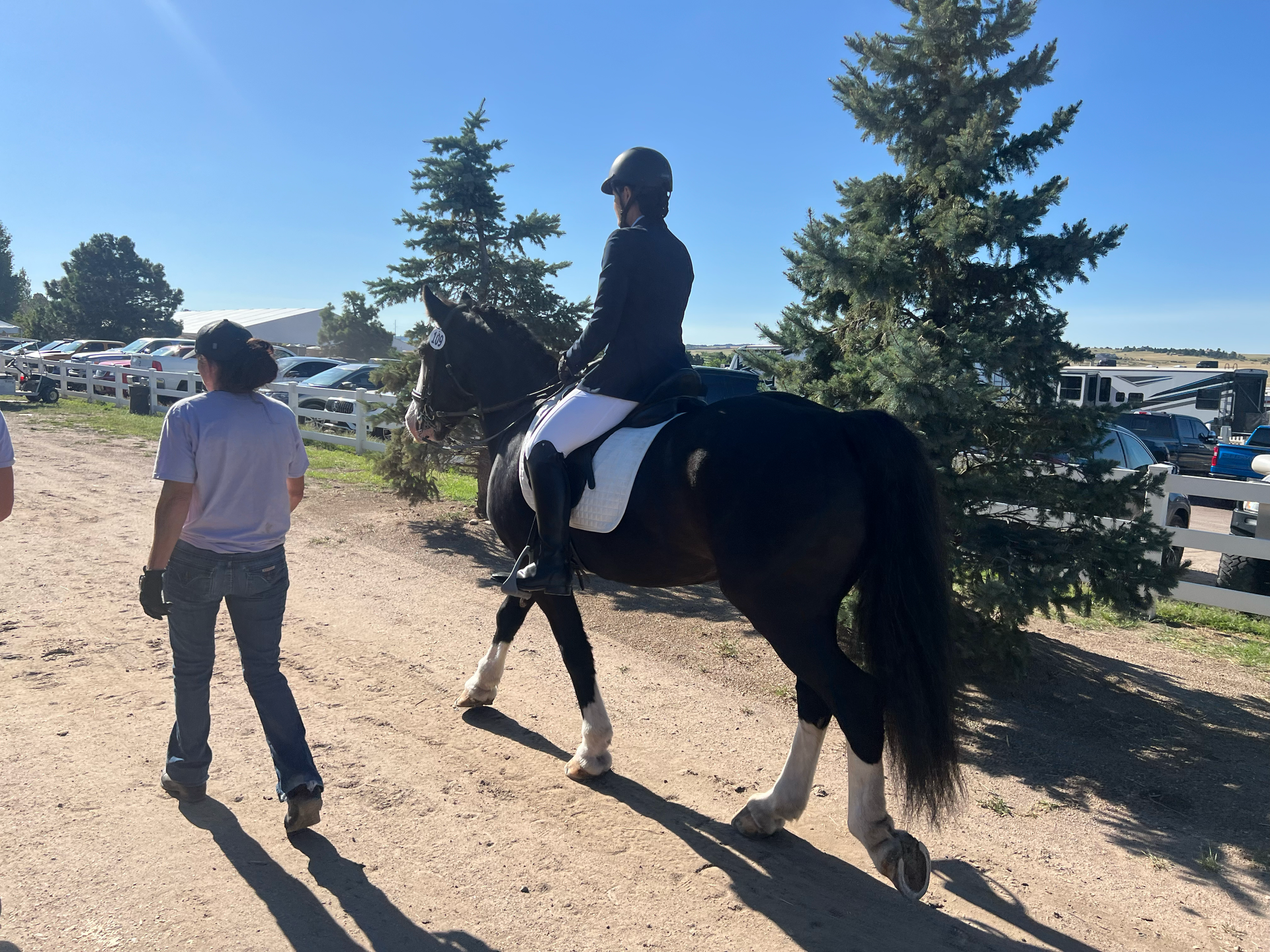 Person riding a black horse beside a guide on a dirt path, with trees and parked vehicles in the background with slow & say hello in colorado