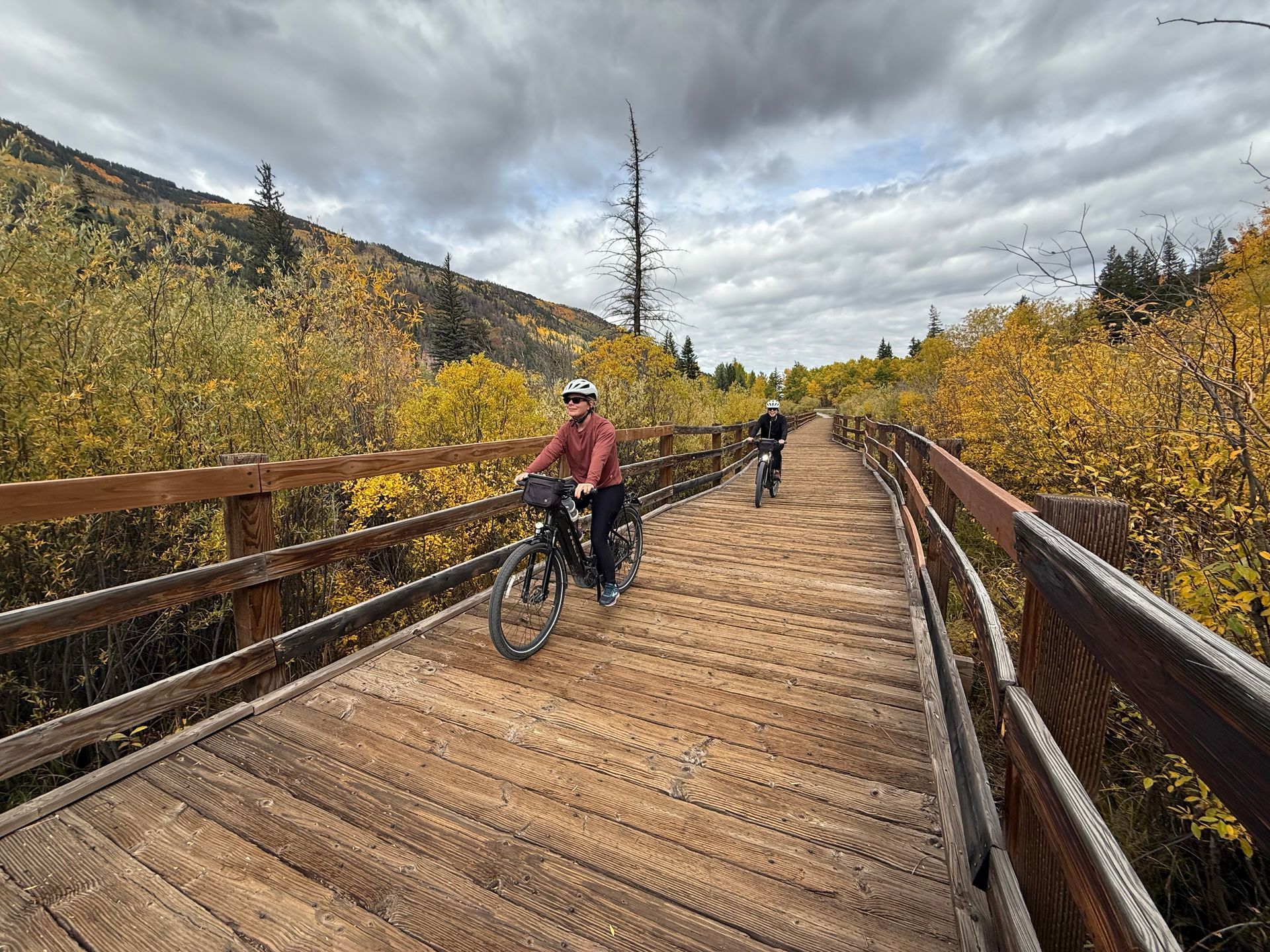 Cyclists ride across a wooden boardwalk through autumn trees under a cloudy sky with slow & say hello in colorado