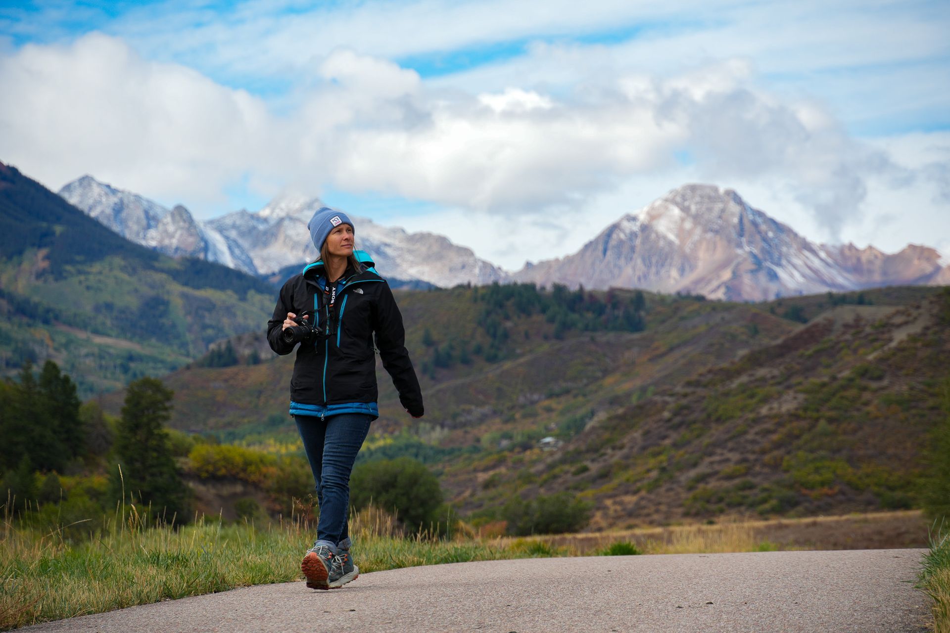 Hiker in black jacket and blue pants walking on a mountain road with snowy peaks and cloudy sky with slow & say hello