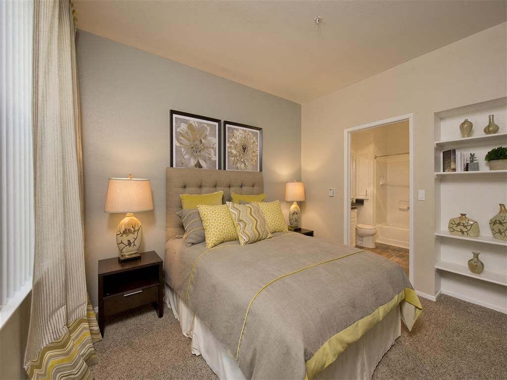 Bedroom in an apartment with a neatly made bed, two bedside lamps, and an open doorway to the bathroom at Rosewalk Apartment Homes in San Jose, CA.