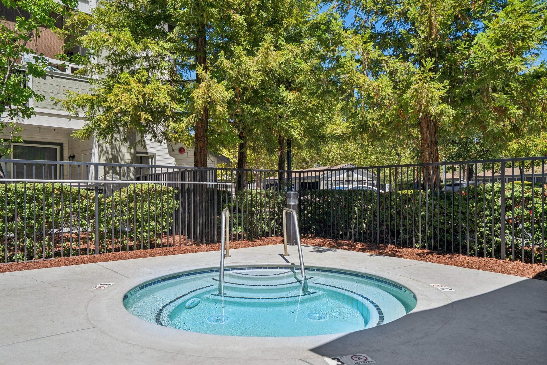 Outdoor courtyard with a small round fountain, black metal fencing, and trees in sunlight