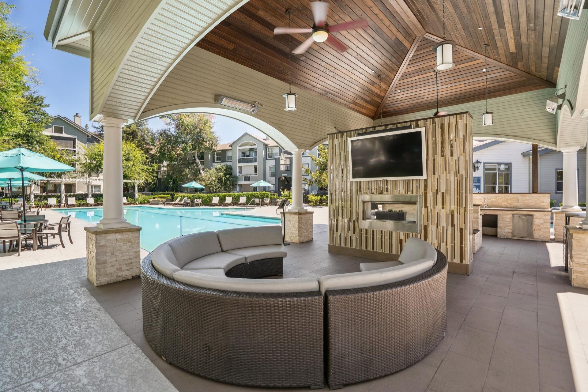 Covered outdoor lounge with circular seating, TV, and ceiling fans by a poolside patio