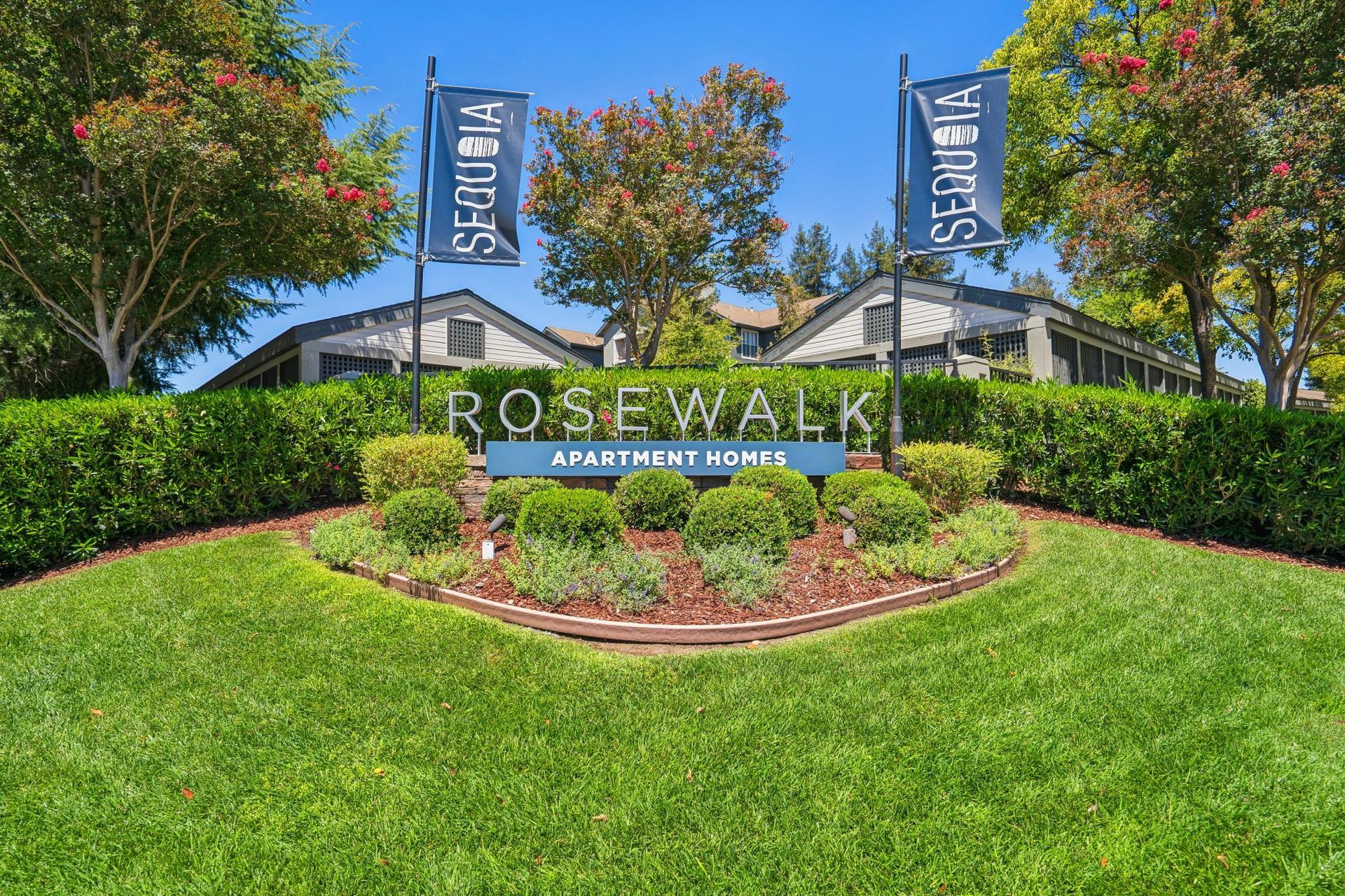 Rosewalk entrance sign amid manicured green lawn and hedges, with blue flags and trees under a clear sky