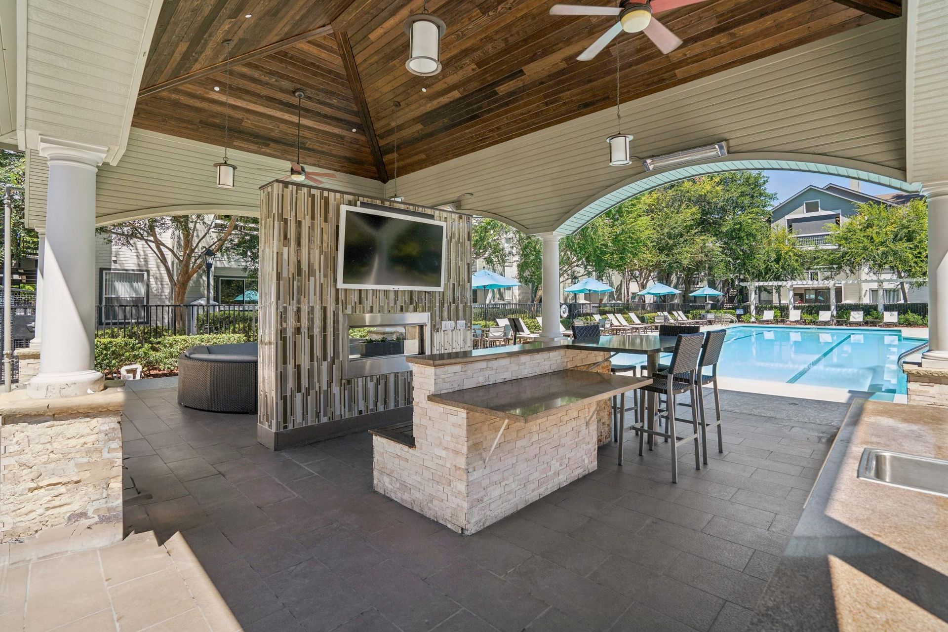 Covered outdoor pool cabana with stone bar, stools, TV, and ceiling fans beside a blue swimming pool.
