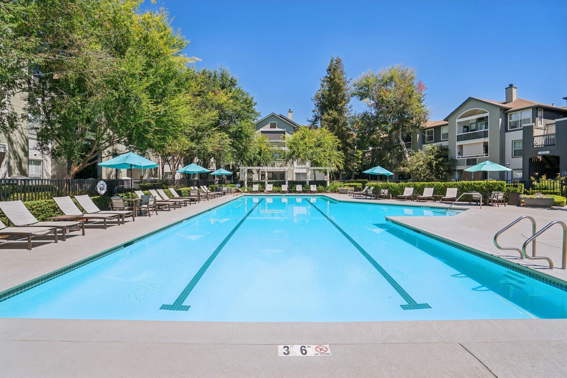 Outdoor swimming pool with clear blue water, lounge chairs, and apartment buildings surrounded by trees