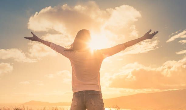 A woman is standing in front of a sunset with her arms outstretched.