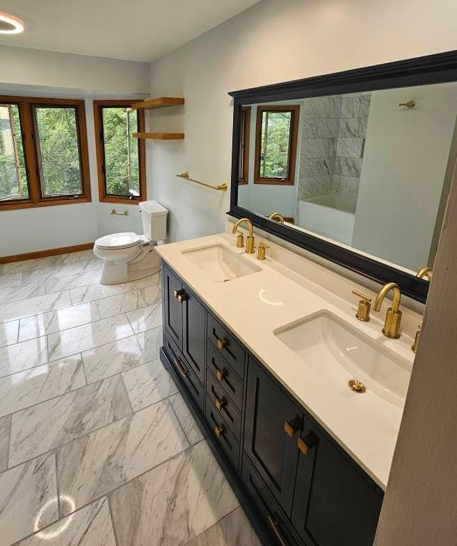 Bright bathroom with black vanity, gold faucets, marble floor, toilet, tub, and windowed walls.
