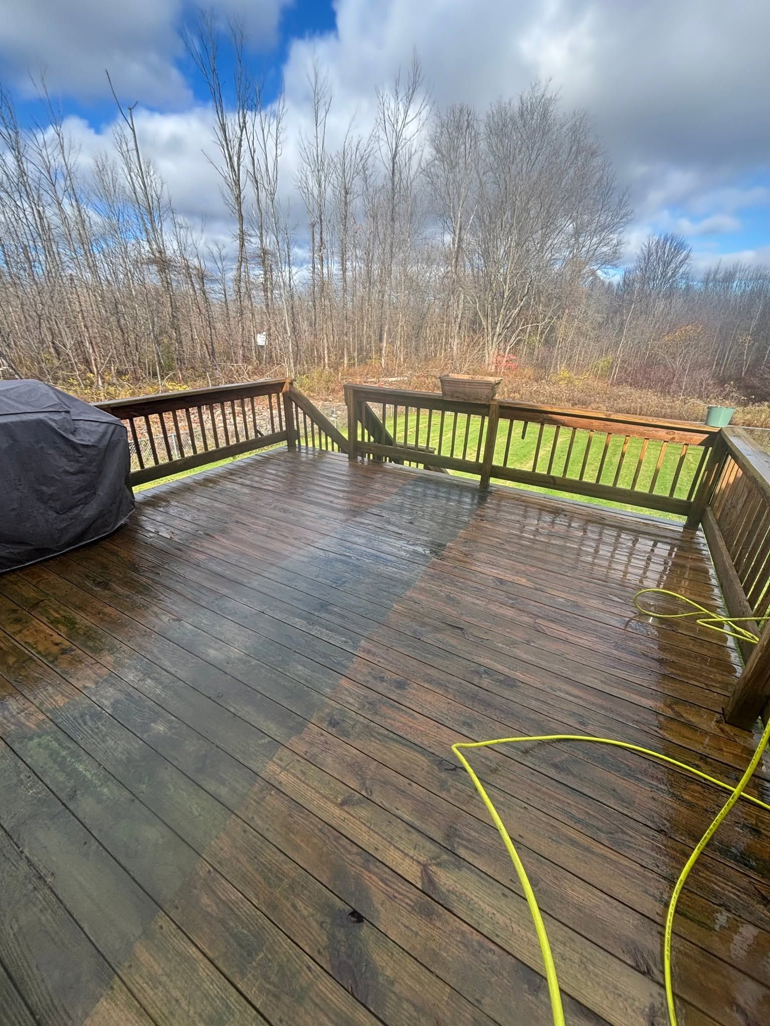 Wet wooden deck with railing, covered grill, and yellow hose overlooking trees under a cloudy sky