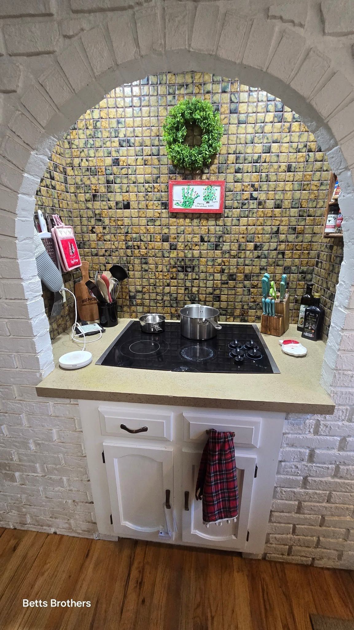 Arched kitchen pass-through with white brick, beige counter, black stovetop, and potted greenery on tiled backsplash.