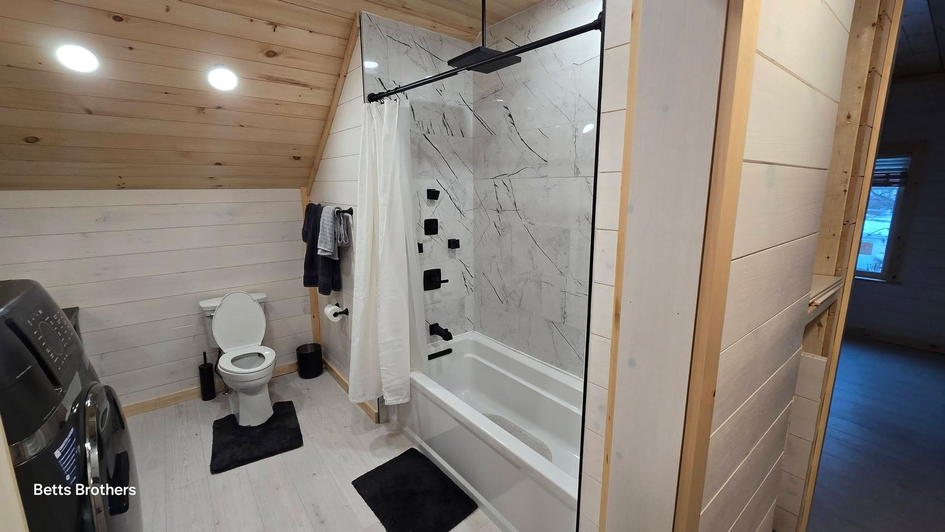 Modern bathroom with wood ceiling, white toilet, glass shower, and black bath mats