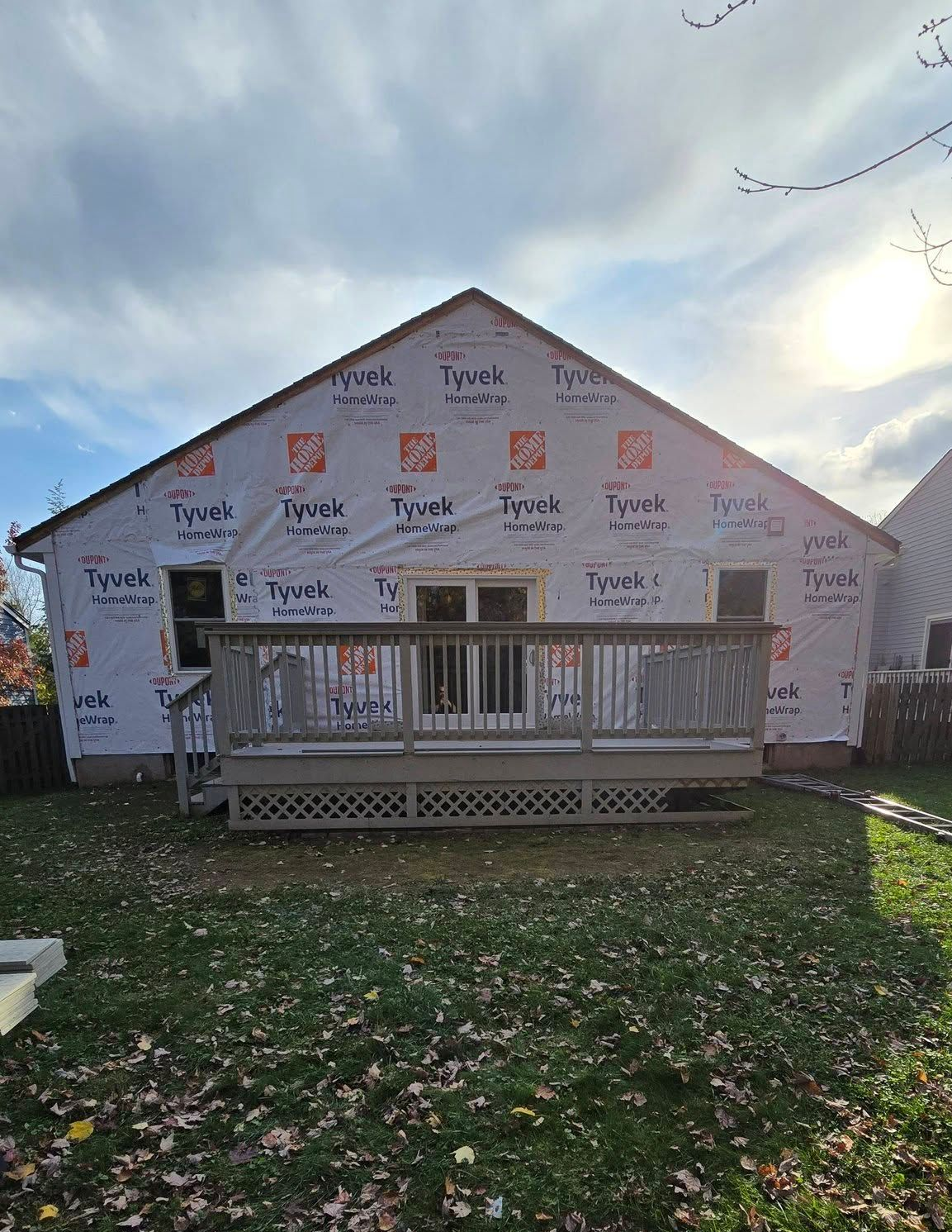 House under construction wrapped in white Tyvek, with a front porch and leaf-covered yard.