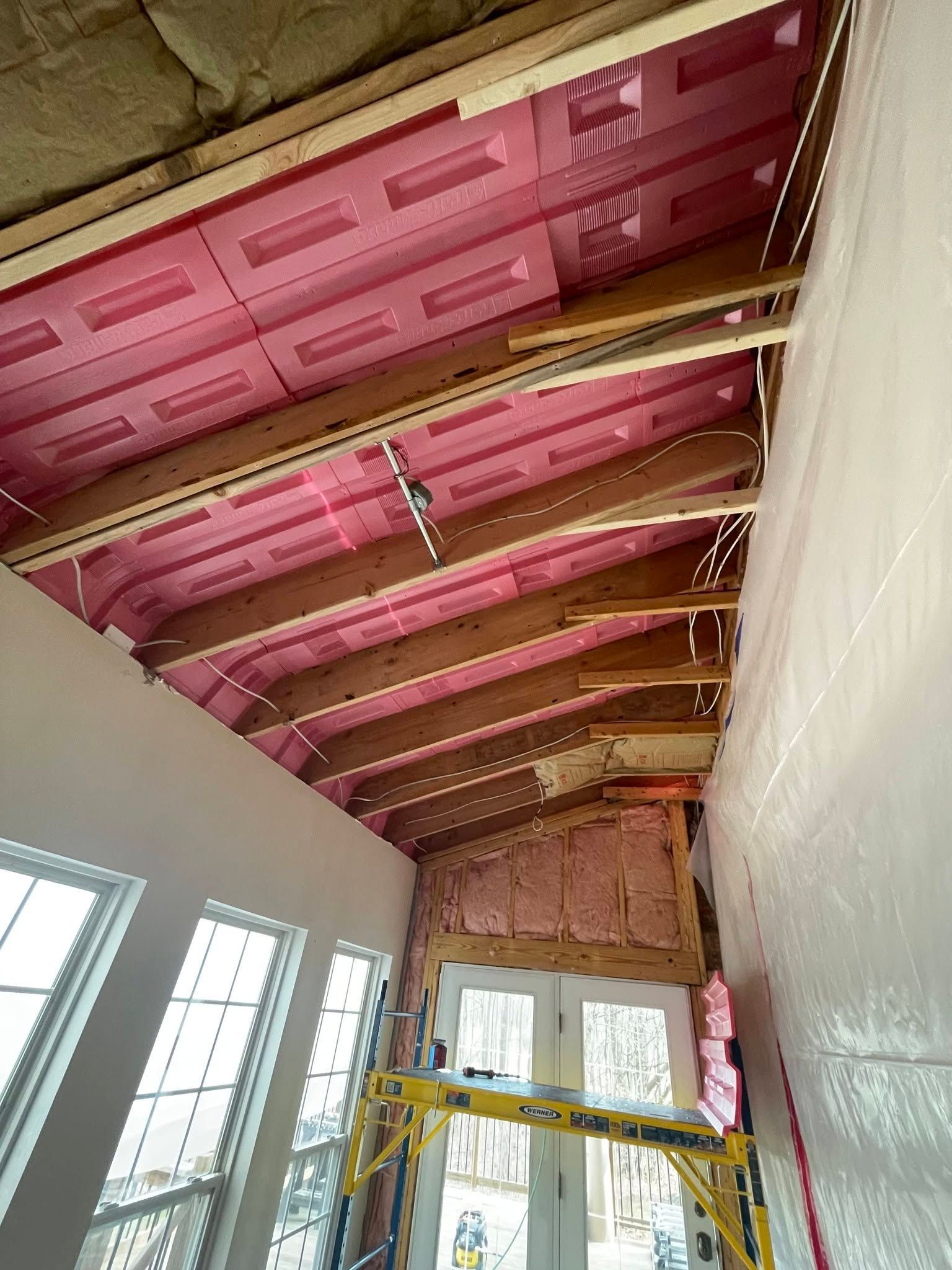Unfinished room with pink insulated ceiling, exposed beams, and windows along one wall.