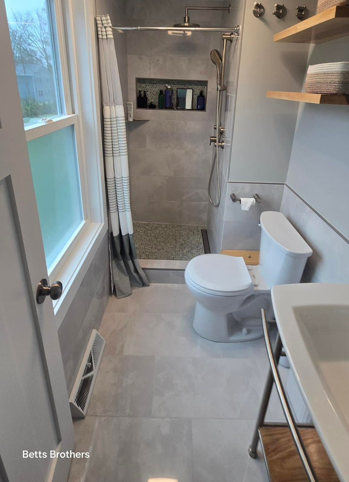 Narrow bathroom with a shower, toilet, sink, window, and shelves, finished in gray and white.