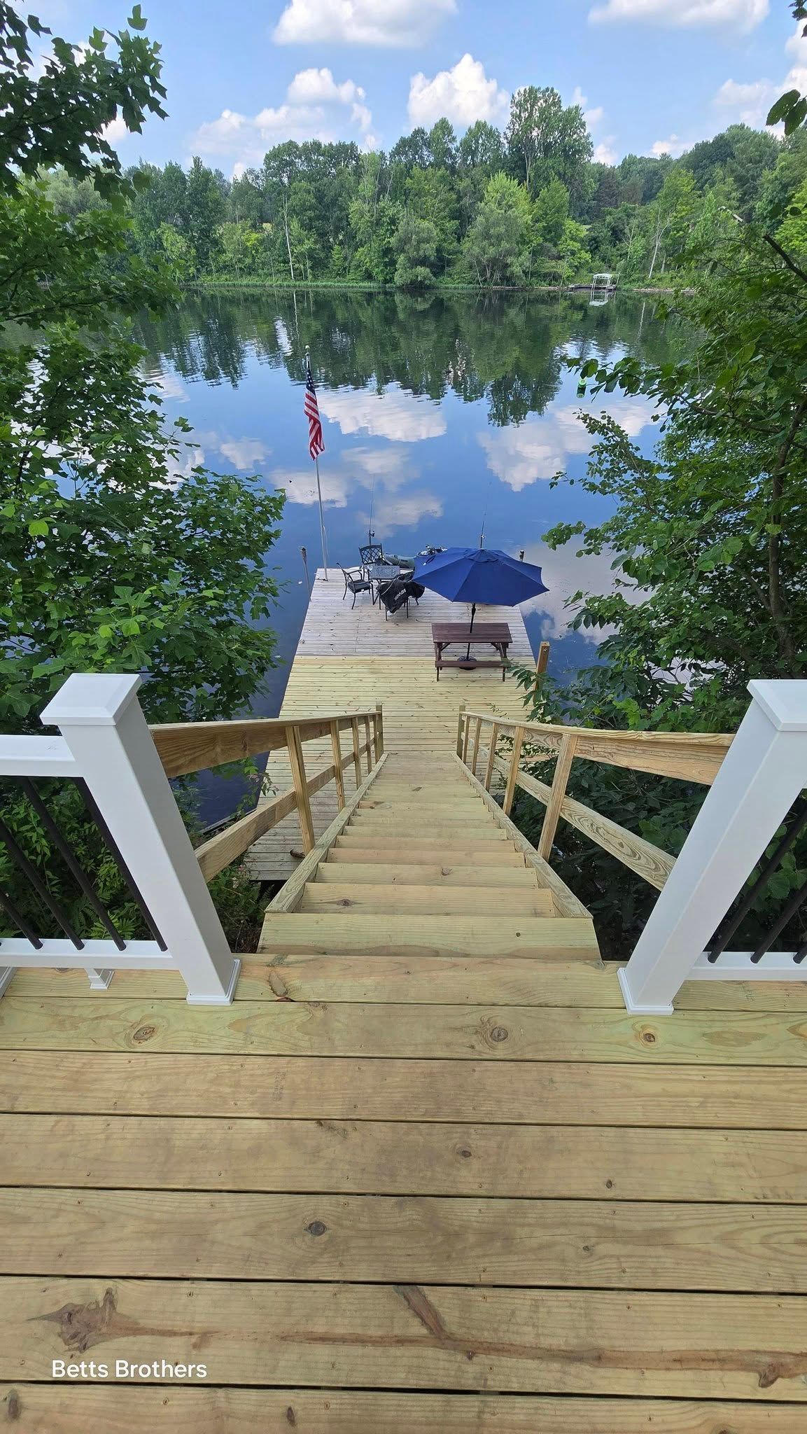 Wooden stairs descend to a lake dock with blue umbrellas, surrounded by green trees and reflected sky.