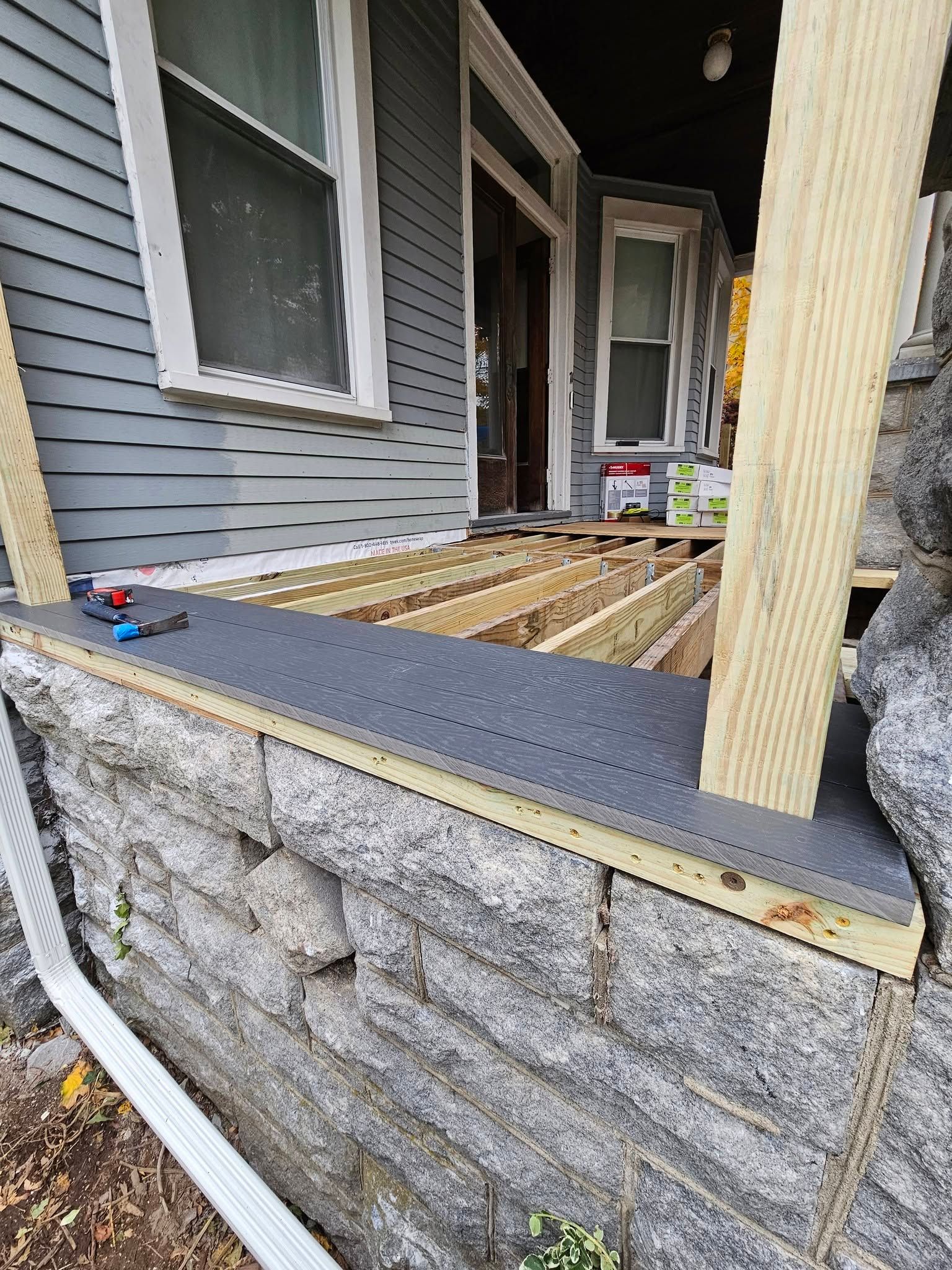 Partially built porch with exposed wooden deck framing beside a gray house and stone foundation