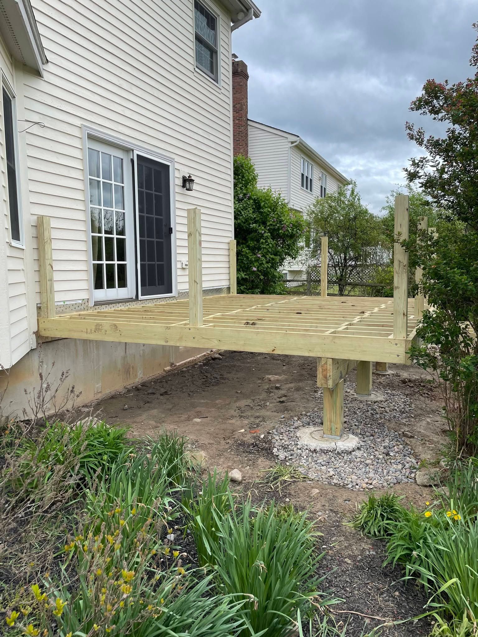Raised wooden deck under construction beside a white house, with posts in gravel and unfinished framing.