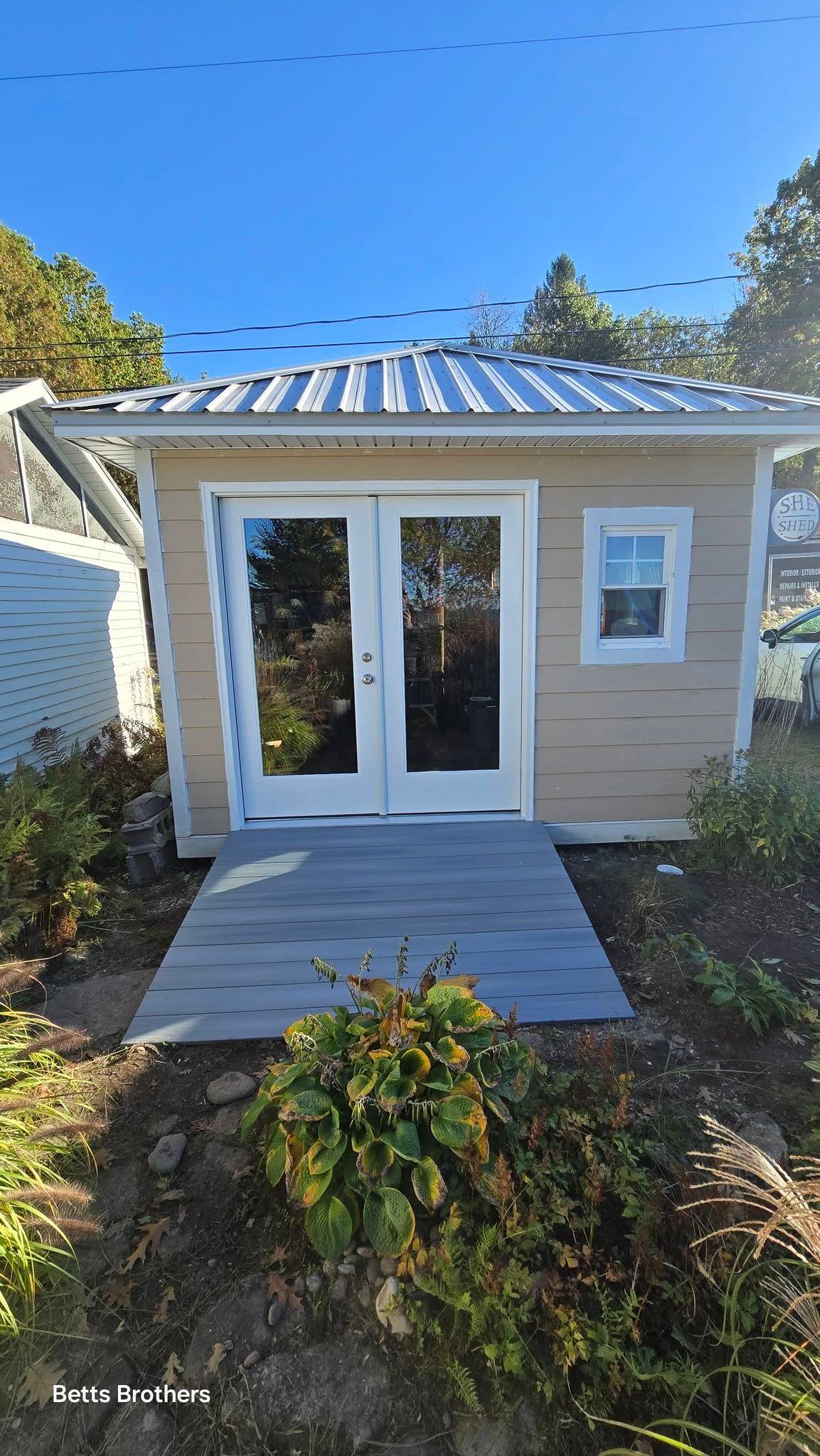 Small beige backyard garden shed with white double doors, a side window, and a short walkway.