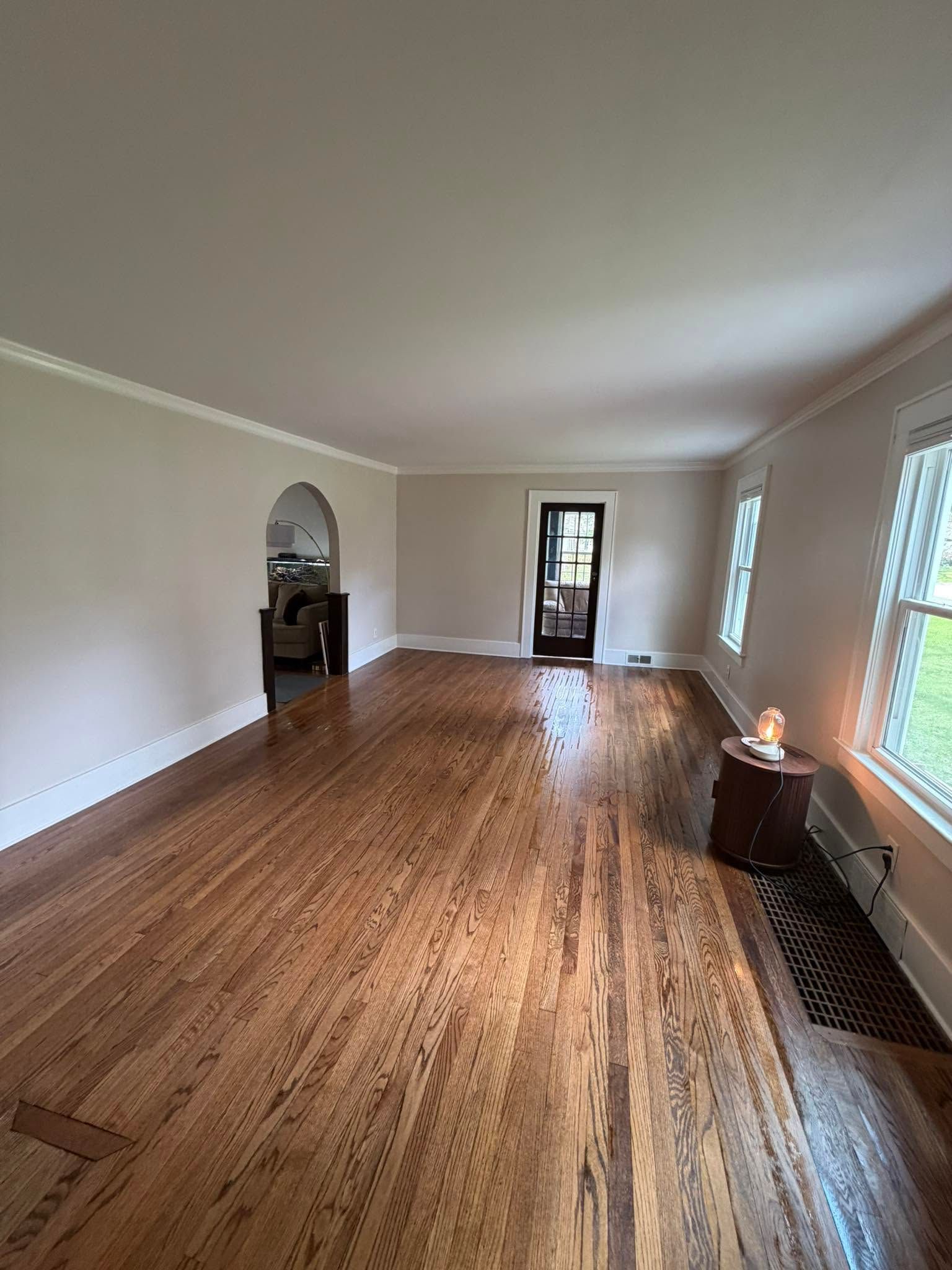 Empty long room with hardwood floors, arched doorway, and bright windows along the right wall.